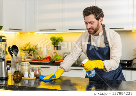 Middle-aged man smiling while cleaning kitchen table with cloth for home hygiene and freshness daily Middle-aged man smiling while cleaning kitchen table with cloth for home hygiene and freshness daily 137994225