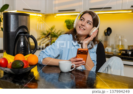 Young woman relaxing in home kitchen with headphones enjoying music while checking smartphone screen Young woman relaxing in home kitchen with headphones enjoying music while checking smartphone screen 137994249