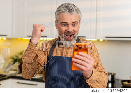 Middle-aged man celebrating big smartphone victory in home kitchen after online success notification 137994262