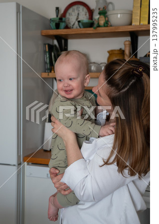 Mother holds smiling baby in kitchen interior with natural light and shelves with home items, warm interaction and everyday domestic moment. Family bonding, motherhood, parenting content 137995235