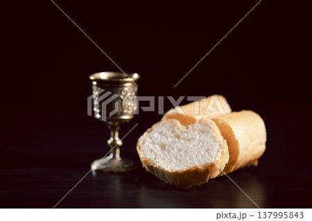 Communion still life with bread on a wooden plate, red wine in a small cup and ornate chalice beside a Holy Bible on dark table, symbolizing Eucharist, worship, faith and remembrance with copy space. 137995843