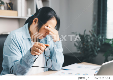 Stressed woman holding eyeglasses and rubbing forehead at home office desk with laptop and documents, burnout and anxiety from deadlines, fatigue, mental health and work stress concept. stress burnout 137995883