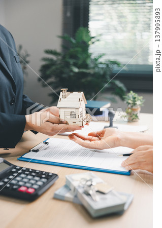 Real estate agent handing wooden house model to client over property contract at office desk, concept of home purchase, mortgage approval, ownership transfer, key handover and investment. 137995893