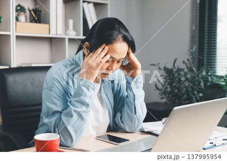 Stressed woman holding head while working on laptop at home office desk, overwhelmed by deadlines and paperwork, burnout, anxiety, fatigue, remote work stress and mental health concept. burnout 137995954