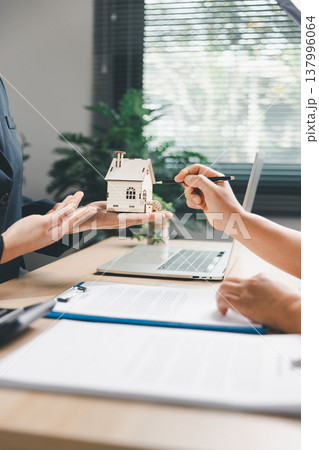 Real estate agent presenting wooden house model to client pointing during property consultation at office desk with laptop and contract, concept of home purchase, mortgage planning and ownership. 137996064