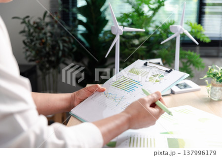 Sustainable business professional reviewing ESG report on clipboard with green charts, wind turbines and solar model on desk, planning renewable energy strategy and carbon reduction for green finance. Sustainable business professional reviewing ESG report on clipboard with green charts, wind turbines and solar model on desk, planning renewable energy strategy and carbon reduction for green finance. 137996179