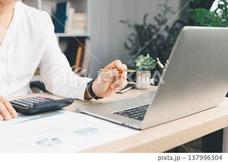 Business analyst reviewing financial report on laptop with calculator and chart documents at office desk, budgeting and accounting for corporate finance, audit, tax and investment planning. financial 137996304