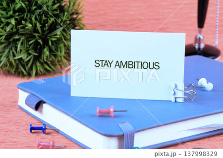 White card shows STAY AMBITIOUS resting on a serene blue journal. Bright pins and a verdant plant adorn the warm wooden desk, encouraging aspirations White card shows STAY AMBITIOUS resting on a serene blue journal. Bright pins and a verdant plant adorn the warm wooden desk, encouraging aspirations 137998329