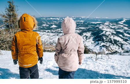Kids standing on top of cliff in winter mountains at sunset and enjoying view of nature Kids standing on top of cliff in winter mountains at sunset and enjoying view of nature 137999310
