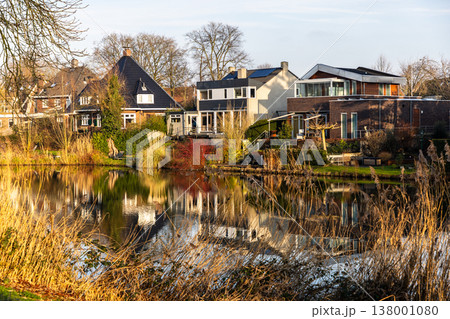 Modern and traditional suburban houses reflected in a calm pond at golden hour. Serene lakeside neighborhood with autumn reeds, sunlit architecture, and peaceful atmosphere in Europe. 138001080