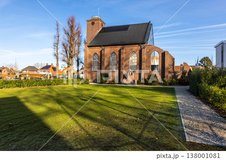 Historic brick church with tall arched windows and a modern roof extension, surrounded by a green lawn, hedges and nearby houses, captured on a sunny day in a peaceful small town. 138001081