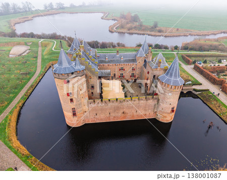 High aerial view of the historic Muiderslot Castle, a perfectly preserved medieval brick fortress near Amsterdam, Netherlands, surrounded by a dark moat and a network of water channels on a misty day. 138001087