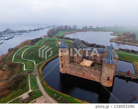 High aerial view of the historic Muiderslot Castle, a perfectly preserved medieval brick fortress near Amsterdam, Netherlands, surrounded by a dark moat and a network of water channels on a misty day. High aerial view of the historic Muiderslot Castle, a perfectly preserved medieval brick fortress near Amsterdam, Netherlands, surrounded by a dark moat and a network of water channels on a misty day. 138001090
