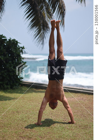 Man holding handstand on grass near ocean in tropical setting Man holding handstand on grass near ocean in tropical setting 138001566