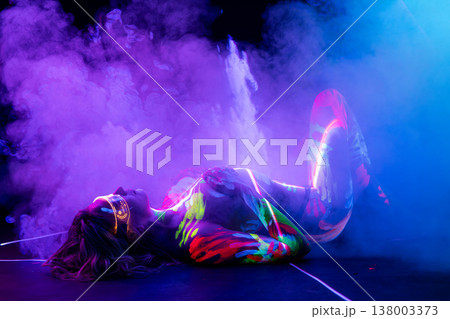 Cyberpunk model in glowing neon body paint and futuristic glasses lying in colorful smoke. 138003373