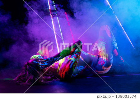Woman in glowing UV body paint lying in smoky, futuristic neon light. 138003374