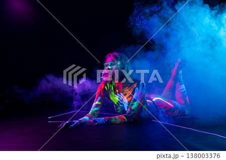 Woman covered in neon UV body paint with glowing glasses lies in colorful smoke. Woman covered in neon UV body paint with glowing glasses lies in colorful smoke. 138003376