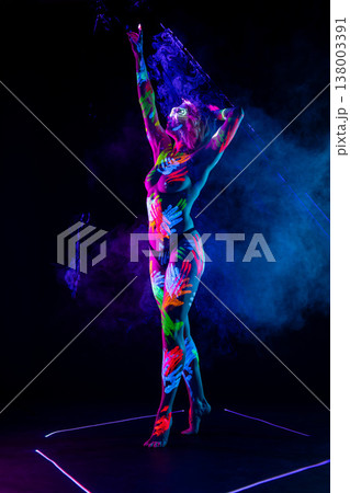 Woman with neon handprint body art and glowing glasses posing in vibrant smoke under UV light. Woman with neon handprint body art and glowing glasses posing in vibrant smoke under UV light. 138003391