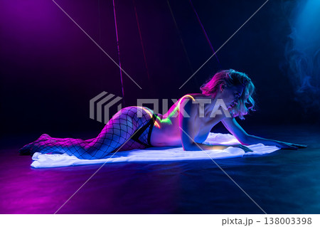 Sensual woman in fishnet stockings lying in a futuristic club setting with neon blue and pink lights, smoke, and red laser beams. 138003398