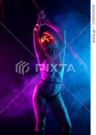 Sensual cyberpunk woman in glowing glasses posing with neon pink and blue laser beams in a dark, smoky studio. Sensual cyberpunk woman in glowing glasses posing with neon pink and blue laser beams in a dark, smoky studio. 138003426