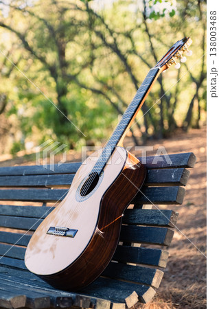 Beautiful classical guitar on a bench in the park with lens flare. Photo of a new wooden guitar with nylon strings outdoors in the summer during sunset. No people. Beautiful string instrument. 138003488