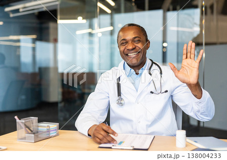 African american male doctor wearing a white lab coat and stethoscope, smiling warmly and waving hand while sitting at a desk in a modern medical office, providing virtual care 138004233