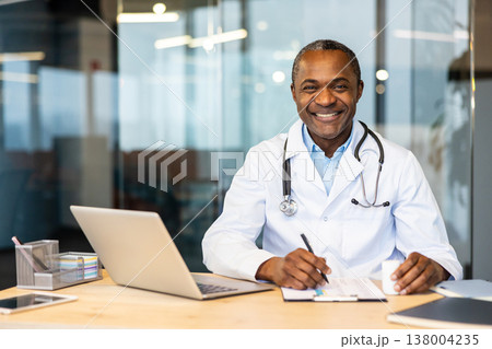 Mature medical doctor in a lab coat and stethoscope, smiling confidently while working at a desk with a laptop and documents, embodying professionalism and expertise in healthcare Mature medical doctor in a lab coat and stethoscope, smiling confidently while working at a desk with a laptop and documents, embodying professionalism and expertise in healthcare 138004235