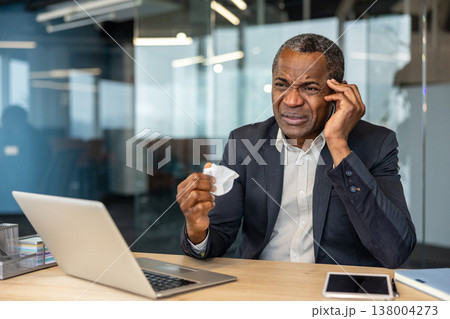 Mature professional man at his office desk rubbing his temple and holding a crumpled tissue, looking unwell and fatigued from headache, stress or flu while working on a laptop 138004273