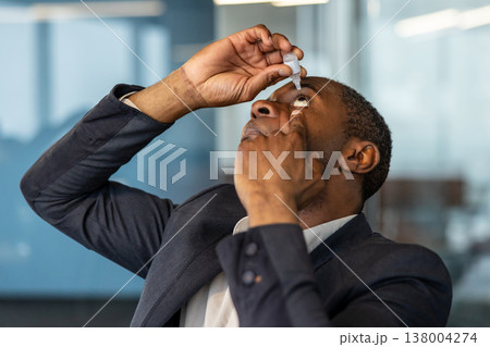 Man in a business suit applying eye drops to his eye, seeking relief from dry eye syndrome or tired vision while maintaining professional focus within a modern office environment Man in a business suit applying eye drops to his eye, seeking relief from dry eye syndrome or tired vision while maintaining professional focus within a modern office environment 138004274