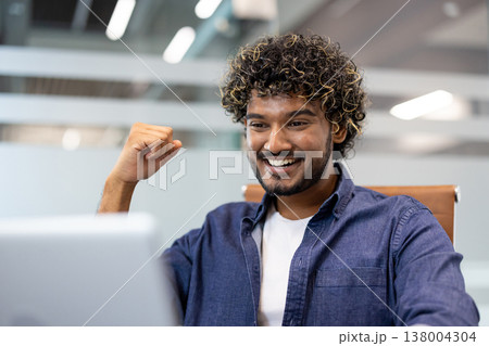 Photo of a young Indian man sitting at his desk and looking at his laptop screen, rejoicing in his success. 138004304