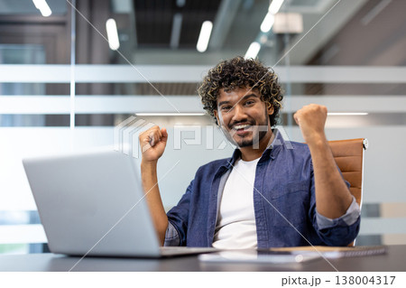 Portrait of a happy young Indian man sitting in the office at a table with a laptop, raising his hands up and rejoicing. Portrait of a happy young Indian man sitting in the office at a table with a laptop, raising his hands up and rejoicing. 138004317