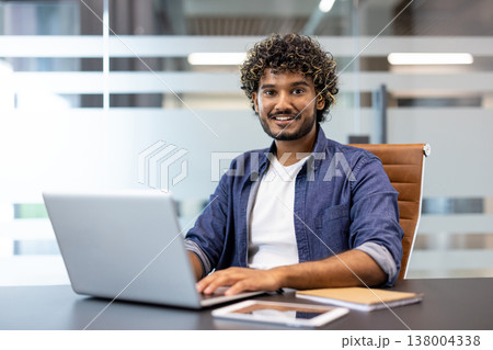 Portrait of a young Indian man sitting at a desk in the office, working on a laptop and smiling, looking at the camera. Portrait of a young Indian man sitting at a desk in the office, working on a laptop and smiling, looking at the camera. 138004338