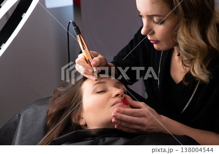 Young woman making tatouage for eyebrows in a dressing makeup room. Eyebrows make up process. Make-up artist in beauty studio doing makeup for beautiful girl. Making mua. 138004544