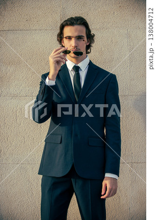 Handsome young man model in a blue elegant business suit poses with sunglasses against a light stone wall outdoors in summer. Male Fashion model poses in a stylish blue suit and white shirt. Handsome young man model in a blue elegant business suit poses with sunglasses against a light stone wall outdoors in summer. Male Fashion model poses in a stylish blue suit and white shirt. 138004712