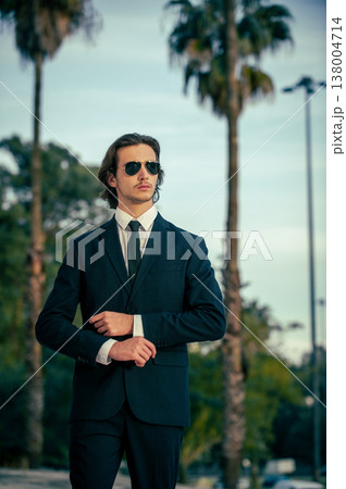Stylish portrait of  a male fashion model with sunglasses in a elegance blue suit with tie. Handsome young man model in modern business suit poses with sunglasses posing in the street. 138004714