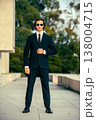 Stylish portrait of  a male fashion model with sunglasses in a elegance blue suit with tie. Handsome young man model in modern business suit poses with sunglasses posing in the street. 138004715