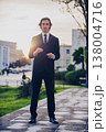 Portrait of  a male fashion model  in a stylish blue suit with tie posing in the city. Handsome young man model in elegant business suit poses outdoors with bright sunlight on background. 138004716