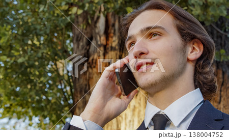 Portrait of the  Businessman in a business suit in the park  with mobile phone.  Smiling businessman talking on a cell phone., outdoors. Young man in a business  suit talking on a smartphone in nature 138004752