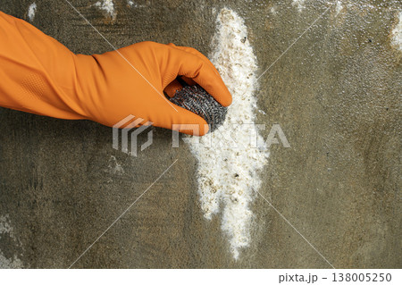Close-Up of Hand Wearing Orange Protective Rubber Glove Using Steel Wool Scrubber to Remove White Mold and Efflorescence From Rough Concrete Wall Surface 138005250
