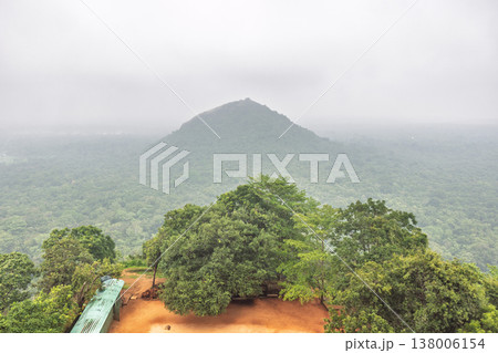 Sigiriya ancient rock fortress in Sri Lanka. Panoramic view of a lush green tropical forest and distant mountain under an overcast, hazy sky, serene landscape 138006154