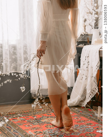 Barefoot woman in sheer dress holding blossom branch in bright airy interior. 138006593