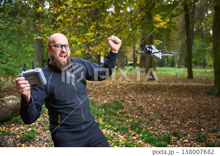 Man celebrates while flying a drone with remote controller. Bearded man operating quadcopter in a park, reacting with excitement during flight, concept of modern technology and hobby Man celebrates while flying a drone with remote controller. Bearded man operating quadcopter in a park, reacting with excitement during flight, concept of modern technology and hobby 138007682