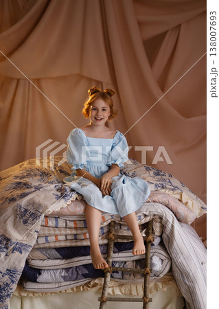 Little girl in blue dress sitting on stacked mattresses like princess and the pea, studio drapery background Little girl in blue dress sitting on stacked mattresses like princess and the pea, studio drapery background 138007693