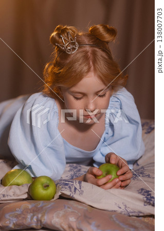 Little girl in blue dress with crown holding green apple, serious portrait, studio drapery Little girl in blue dress with crown holding green apple, serious portrait, studio drapery 138007703