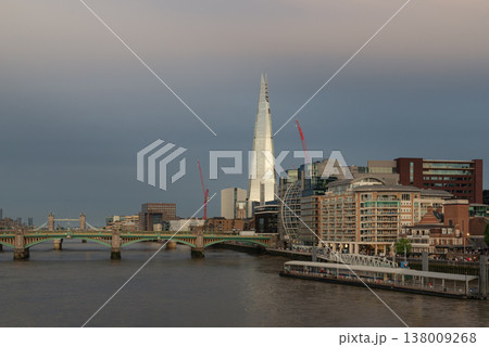 Beneath a soft London sky, The Shard stands as a luminous needle of glass, piercing the skyline with its celebrated pyramidal form. In the foreground, the steady waters of the Thames are spanned by 138009268