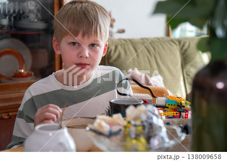 Boy looks at camera while eating at table with sweets, toy in home interior with natural light, everyday meal moment and interaction in domestic setting. Childhood activity, family life, daily routine Boy looks at camera while eating at table with sweets, toy in home interior with natural light, everyday meal moment and interaction in domestic setting. Childhood activity, family life, daily routine 138009658