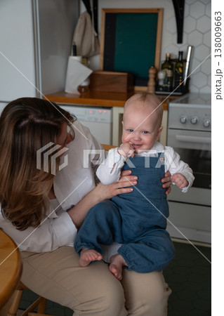 Mother sits with smiling baby in kitchen interior with natural light and home setting, close interaction and everyday family moment in domestic environment. Family bonding, motherhood, parenting Mother sits with smiling baby in kitchen interior with natural light and home setting, close interaction and everyday family moment in domestic environment. Family bonding, motherhood, parenting 138009663