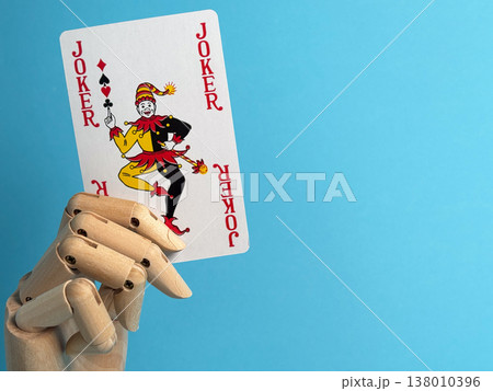Wooden hand holding a joker playing card in front of a blue background indoors 138010396