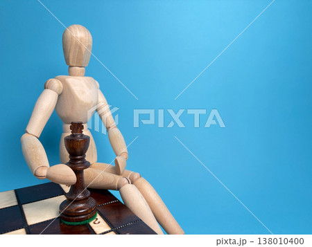 Wooden figure sits on a chessboard with a king piece in front against a blue background showing a game in progress 138010400