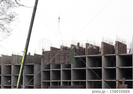 Heavy crane lifting a large formwork panel at a construction site. Unfinished concrete building structure with exposed rebar and workers. Urban development and industrial engineering. Copy space Heavy crane lifting a large formwork panel at a construction site. Unfinished concrete building structure with exposed rebar and workers. Urban development and industrial engineering. Copy space 138010889